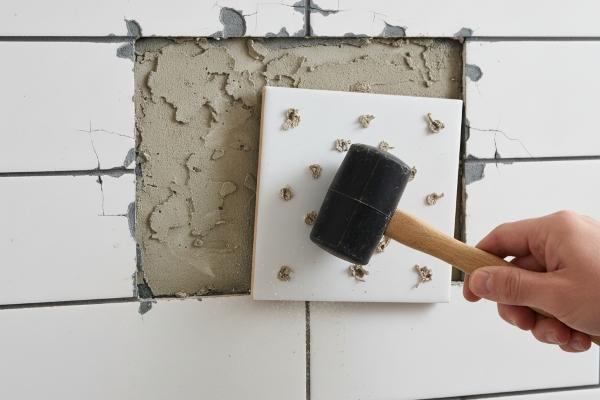 Hollow-sounding subway tile being tapped with wooden handle showing poor adhesive coverage underneath
