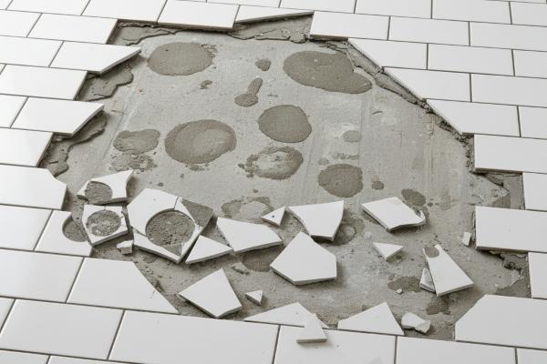 Close-up of a square white bathroom floor tile that is visibly loose or lifting, revealing a hollow space underneath due to poor adhesive coverage