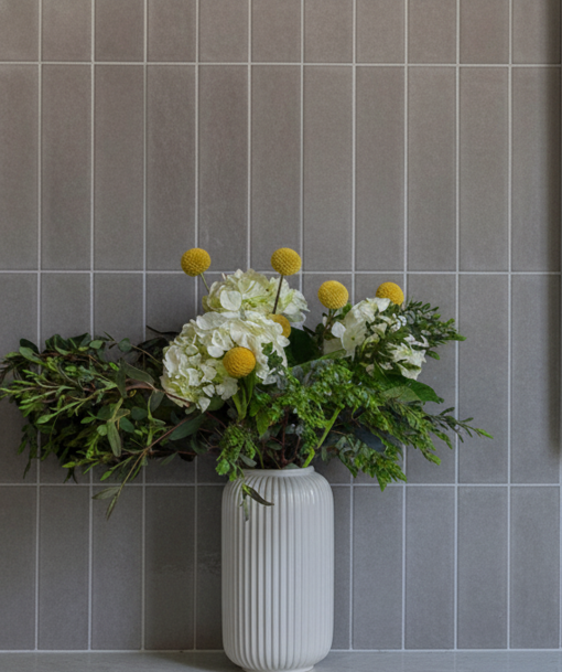 A white, ribbed vase holding a bouquet of white hydrangeas, yellow billy buttons, and green foliage against a grey vertical subway tile wall.
