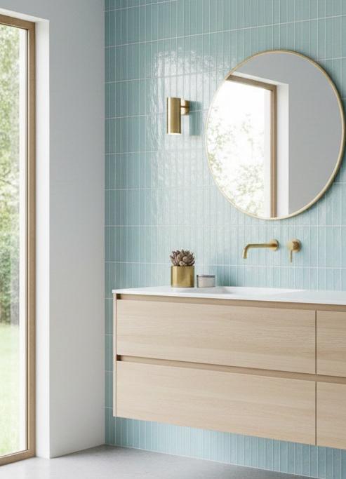 A modern Scandinavian-style bathroom featuring a light wood floating vanity and a round gold-rimmed mirror set against a wall of pale blue, vertically stacked subway tiles.
