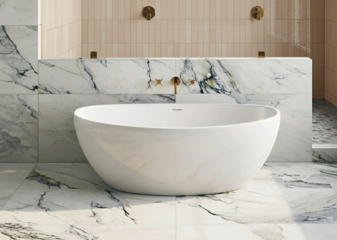sleek white freestanding oval bathtub set against bold marble-look porcelain wall and floor tiles, complemented by gold fixtures and vertical beige subway tiles.