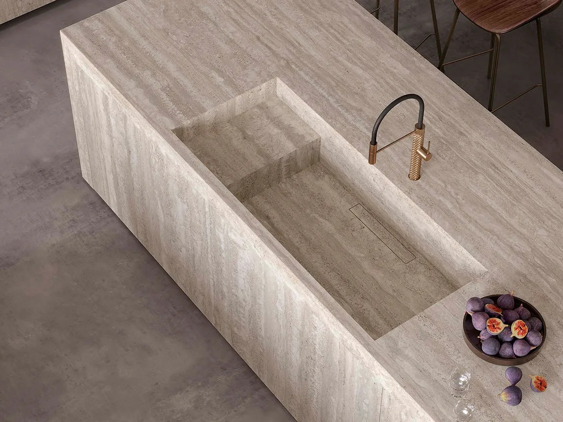 Travertino Chiaro porcelain slabs crafted into waterfall island with carved sink basin and fresh figs.