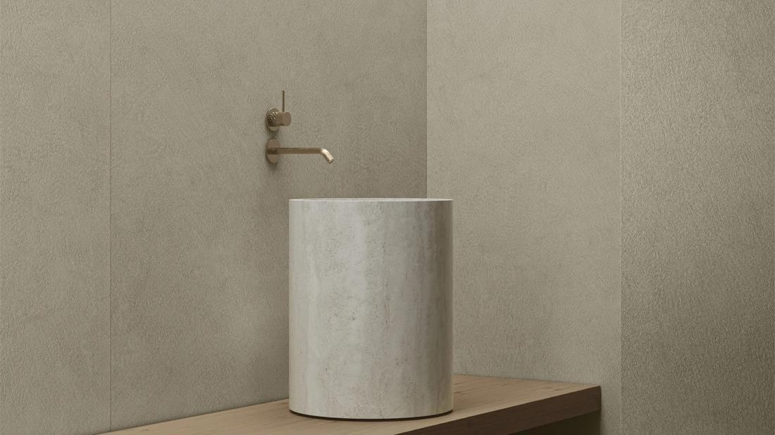 Plaster Bone porcelain slabs bathroom walls with cylindrical sink and bronze wall faucet.